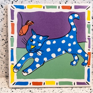 May include: A blue and white spotted cat with a yellow nose and whiskers chases a red fish on a purple, green, and yellow background. The cat is outlined in black and the fish is outlined in black. The background is a colourful tile with a border.