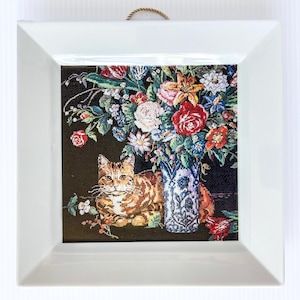 May include: A framed needlepoint featuring a tabby cat resting near a vase of colorful flowers. The flowers include red roses, blue and white daisies, and other blooms. The frame is white and square.