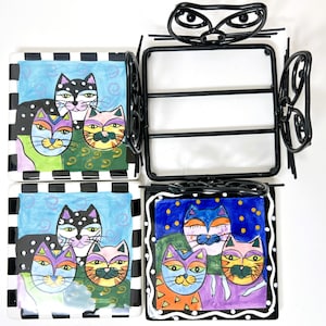 May include: A decorative cat-themed napkin holder with four square ceramic coasters. Each coaster features a colorful, whimsical cat design with black and white checkered borders. The holder is black metal with a cat face design.
