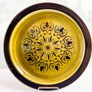 Pode incluir: Um prato de servir grande, redondo e de cerâmica com uma borda marrom escura e um centro amarelo-verde. O centro apresenta um desenho floral preto e marrom.
