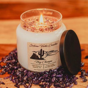 Può includere: Una candela Nag Champa in cera di soia bianca accesa in un barattolo di vetro con coperchio nero. La candela ha un'etichetta con un paesaggio desertico e testo. La candela è circondata da pietre preziose viola.