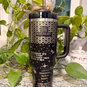 Puede incluir: Un vaso Stanley negro y plateado con asa y pajita. El vaso presenta el logotipo de Stanley y un patr&oacute;n decorativo. El cuerpo del vaso tiene texto e ilustraciones en blanco. El texto incluye frases como "Rider without their dragon is dead."