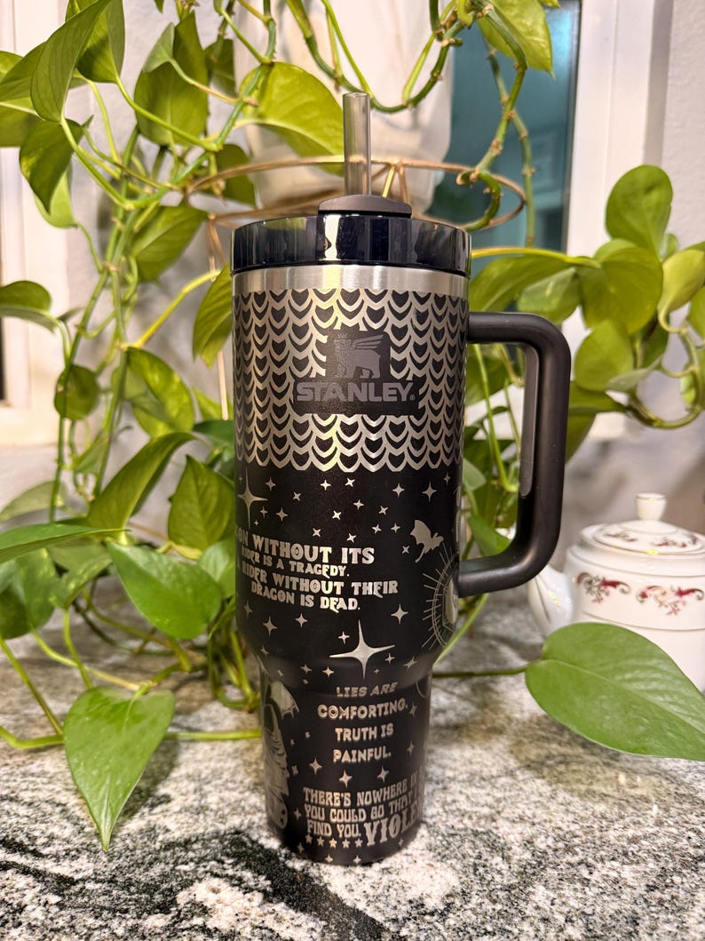 Puede incluir: Un vaso Stanley negro y plateado con asa y pajita. El vaso presenta el logotipo de Stanley y un patr&oacute;n decorativo. El cuerpo del vaso tiene texto e ilustraciones en blanco. El texto incluye frases como "Rider without their dragon is dead."