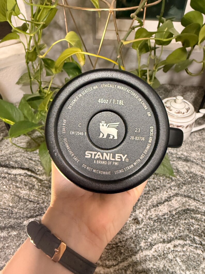 Puede incluir: Taza de viaje negra de la marca Stanley, 1,18L, con asa. La base de la taza es visible, mostrando el logotipo de la marca y el texto: "Designed in Seattle WA, Ethically Manufactured in China-AM, Dishwasher Safe, Do Not Microwave."