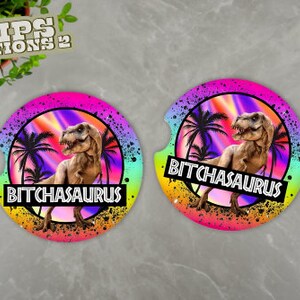 May include: Two round, colorful coasters with a dinosaur silhouette and the text "BITCHASAURUS". The coasters have a rainbow gradient background with palm trees and a black splatter effect.