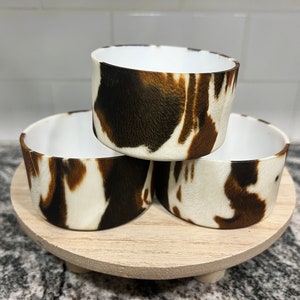 May include: Three small white ceramic bowls with a brown and white cow print design are stacked on a wooden tiered stand.