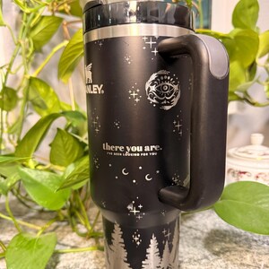 Puede incluir: Taza de viaje Stanley negra con borde, asa y base plateados. La taza presenta un dise&ntilde;o de bosque plateado en la base, con estrellas y un patr&oacute;n de media luna. El texto "there you are. I've been looking for you" est&aacute; impreso.