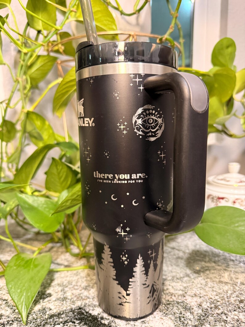 Puede incluir: Taza de viaje Stanley negra con borde, asa y base plateados. La taza presenta un dise&ntilde;o de bosque plateado en la base, con estrellas y un patr&oacute;n de media luna. El texto "there you are. I've been looking for you" est&aacute; impreso.