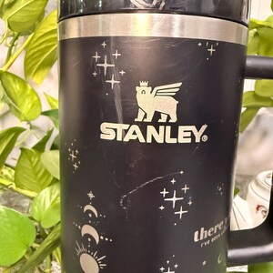 Puede incluir: Taza de viaje Stanley negra con asa, con un logotipo de oso con alas y el nombre de la marca. La taza tiene un dise&ntilde;o celestial con estrellas, lunas y el texto "there I've been".