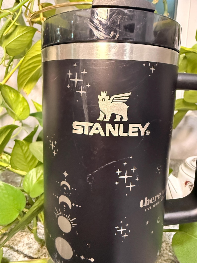 Puede incluir: Taza de viaje Stanley negra con asa, con un logotipo de oso con alas y el nombre de la marca. La taza tiene un dise&ntilde;o celestial con estrellas, lunas y el texto "there I've been".