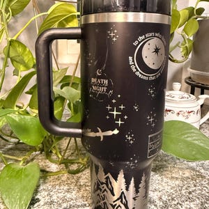Puede incluir: Vaso t&eacute;rmico negro con asa, con dise&ntilde;os plateados grabados de un bosque, estrellas y texto. El texto incluye "to the stars who listen and the dreams that are answered" y "Death, Night, Twilight, Forever".