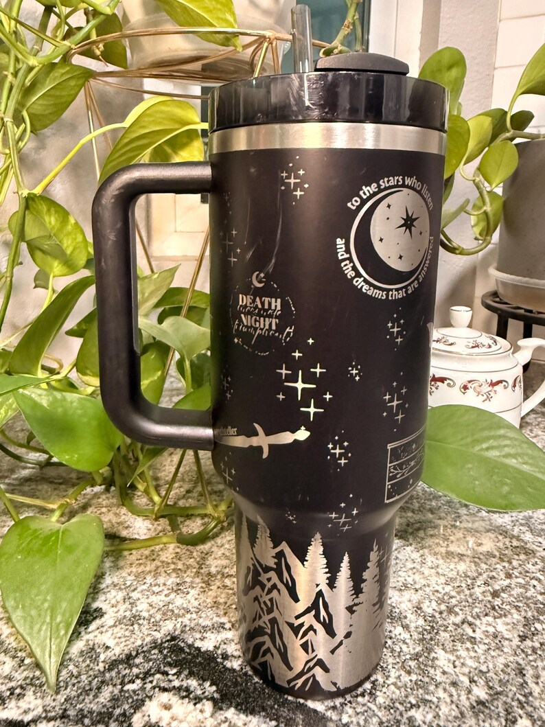 Puede incluir: Vaso t&eacute;rmico negro con asa, con dise&ntilde;os plateados grabados de un bosque, estrellas y texto. El texto incluye "to the stars who listen and the dreams that are answered" y "Death, Night, Twilight, Forever".