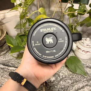 Puede incluir: Taza de viaje negra de la marca Stanley con asa, con el logotipo y el texto de la marca. La taza es de 1,18L y es apta para lavavajillas. La taza la sostiene una persona que lleva un reloj negro con hebilla dorada.