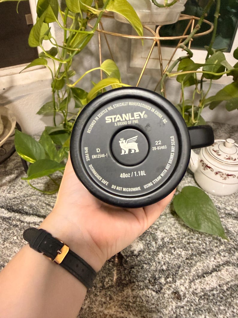 Puede incluir: Taza de viaje negra de la marca Stanley con asa, con el logotipo y el texto de la marca. La taza es de 1,18L y es apta para lavavajillas. La taza la sostiene una persona que lleva un reloj negro con hebilla dorada.