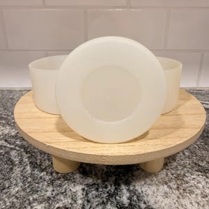 May include: A small, round, light-colored wooden tray with three short, rounded legs. Three white, cylindrical containers and a white, circular lid are arranged on the tray. The background is a white tiled wall and a speckled countertop.