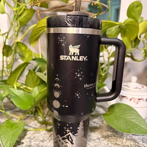 Puede incluir: Vaso Stanley negro con asa y pajita. El vaso presenta un dise&ntilde;o de monta&ntilde;a y fases lunares en plata. El logotipo de Stanley es visible. El texto "there you've been" est&aacute; impreso.