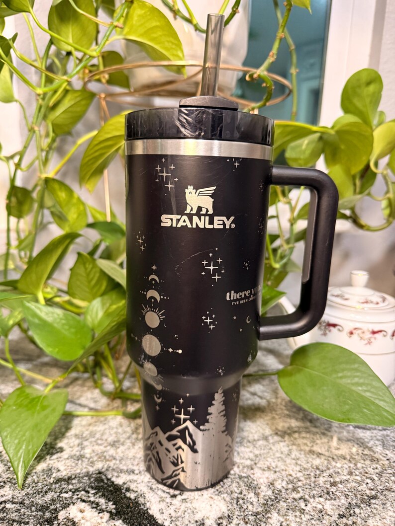 Puede incluir: Vaso Stanley negro con asa y pajita. El vaso presenta un dise&ntilde;o de monta&ntilde;a y fases lunares en plata. El logotipo de Stanley es visible. El texto "there you've been" est&aacute; impreso.