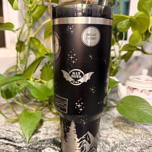 Puede incluir: Vaso negro con detalles plateados y pajita transparente. El vaso presenta el texto "NIGHT COURT" y "BAT BOYS ONLY", junto con un dise&ntilde;o de monta&ntilde;a y &aacute;rboles. Decorado con patrones de estrellas y cruces.
