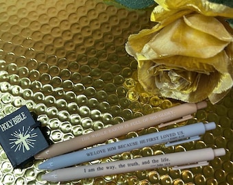 May include: Set of three pens with inspirational bible verses printed on them. The pens are beige, light blue, and gray. The pens are arranged on a gold background with a small black book that says "Holy Bible" on the cover.