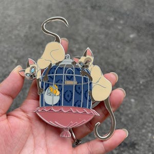 May include: A metal enamel pin featuring two Siamese cats with cream-colored fur and dark brown points, perched on top of a blue birdcage with a dark blue laurel pattern. Inside the cage, a small yellow bird sits within a white eggshell base.