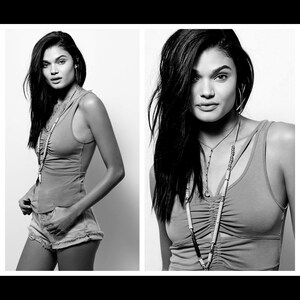 May include: A monochrome image of a woman in a sleeveless top and denim shorts. She is wearing a long necklace and hoop earrings. The woman has long, dark hair and is looking directly at the camera.