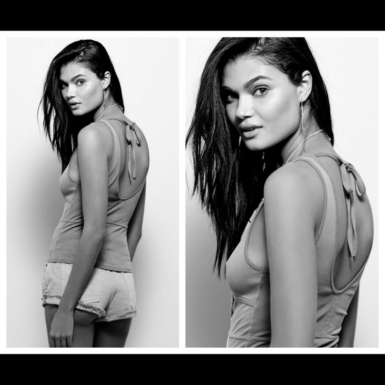 May include: Monochrome image of a woman in a light-coloured vest top with a tie detail at the back and denim shorts. The photograph is a studio shot with a white backdrop.