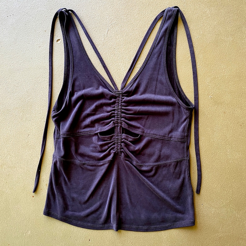 May include: Dark purple tank top with a ruched front and tie straps. The sleeveless top has a V-neckline and two horizontal seams across the front. The straps extend from the shoulders and tie at the back. The fabric has a slight sheen.