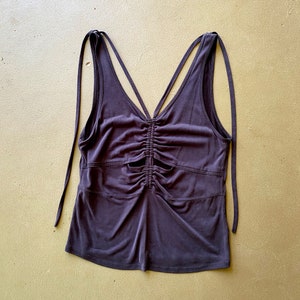 May include: A dark purple tank top with a V-neck and spaghetti straps. The top has a ruched design in the centre and cut-out details. The straps are long and can be tied.