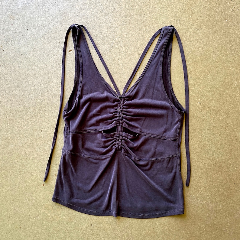 May include: A dark purple tank top with a V-neck and spaghetti straps. The top has a ruched design in the centre and cut-out details. The straps are long and can be tied.
