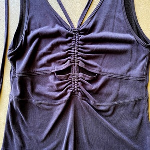 May include: A dark purple tank top with a ruched front and a V-neck design. The top features a criss-cross strap detail in the back and a fitted silhouette. The fabric appears to be a soft, stretchy material.
