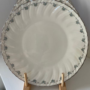 May include: A set of six white ceramic dinner plates with a delicate blue and green floral pattern around the rim. The plates are stacked and displayed on a wooden stand.