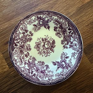 May include: A round ceramic saucer featuring a cream-colored base with a dark purple floral transferware pattern. The design includes a central bouquet of flowers surrounded by a border of intricate blossoms and a dotted edge trim detail.