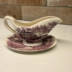 May include: A white ceramic gravy boat and saucer set with a maroon transferware design. The pattern depicts a landscape with buildings, trees, and water. The gravy boat has a handle and a spout. The saucer is round.