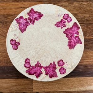 May include: Round, off-white decorative plate with a floral design. The plate features a pattern of magenta-colored leaves and small flower clusters around the rim. The leaves have detailed veining, and the plate has a slightly textured surface.