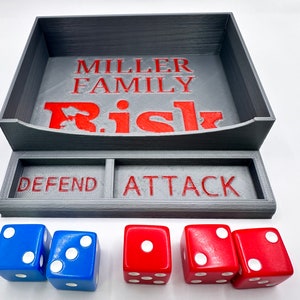 May include: A gray 3D printed Risk game tray with the words "MILLER FAMILY Risk" on the top and "DEFEND" and "ATTACK" on the bottom. There are five dice on the bottom, three red and two blue.