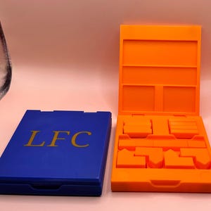 May include: A blue rectangular box with the gold letters "LFC" on the lid is next to an open orange box. The orange box contains several geometric shapes and compartments. The boxes appear to be made of plastic.