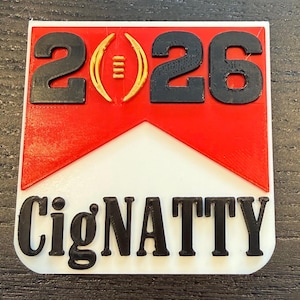 Könnte beinhalten: Ein rechteckiges, mehrfarbiges 3D-gedrucktes Objekt mit dem Text "2026" in Schwarz, einer goldenen Football-Grafik und dem Wort "CigNATTY" in Schwarz. Der Hintergrund weist ein rot-weißes Design auf.