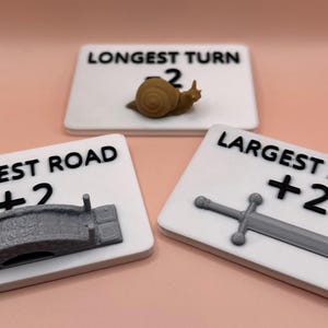 May include: Three white game score markers with black text and 3D printed icons. The first marker says "Longest Road +2" and has a gray bridge icon. The second marker says "Longest Turn +2" and has a brown snail icon. The third marker says "Largest Army +2" and has a gray sword icon.