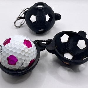 Peut inclure: Un porte-balles de golf noir avec une balle de golf blanche à l'intérieur. Le porte-balles est en forme de ballon de football et possède une petite boucle métallique pour l'attacher à un sac. La balle de golf a des marques roses et le texte "Tour" dessus.