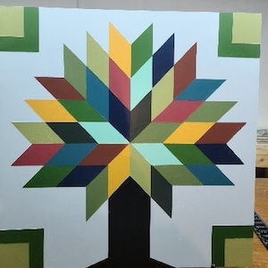 May include: A colorful quilt block with a starburst pattern in shades of green, blue, yellow, red, and brown. The center of the starburst is a dark brown, and the background is a light blue. The block is surrounded by four green squares.