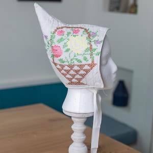 May include: White bonnet with a cross-stitched floral design featuring pink, yellow, and purple flowers in a brown basket. The bonnet is displayed on a white mannequin head and stand, with white ties.