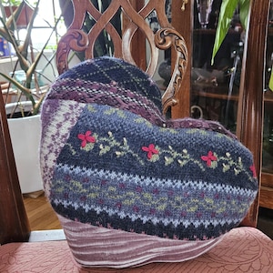 May include: A heart-shaped pillow with a knitted design. The pillow features a mix of colors, including navy blue, purple, and white, with floral embroidery. The base has a striped pattern in shades of pink and purple.