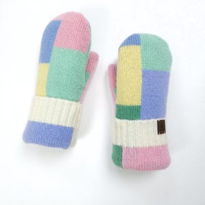 Può includere: Un paio di guanti colorati con un design patchwork. I guanti presentano quadrati rosa, blu, verdi e gialli. I polsini sono bianchi con finiture blu. Una piccola etichetta marrone è su un guanto.