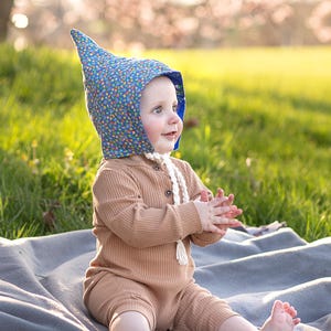 May include: A baby wearing a brown ribbed romper and a blue floral bonnet with a pointed top, sitting on a gray blanket in a grassy field. The bonnet has a blue lining and a white tie.