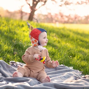 May include: A baby wearing a colorful patchwork bonnet and a tan ribbed jumpsuit sits on a gray blanket in a grassy field. The baby holds a yellow and red toy. A tree with pink blossoms is in the background.
