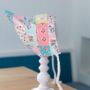 May include: A patchwork baby bonnet with a whimsical print of cherries, mice, and bunnies in pink, blue, and yellow. The bonnet is displayed on a white stand with a braided tie.