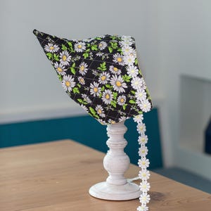 May include: A black bonnet with a white daisy floral pattern and a white daisy trim. The bonnet is displayed on a white decorative stand. The bonnet is quilted with a diamond pattern.