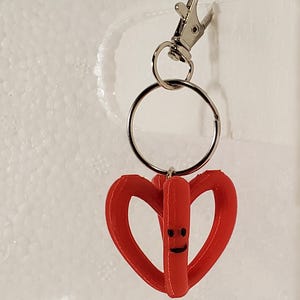 May include: A red heart-shaped keychain with a smiling face, attached to a silver keyring and a lobster clasp. The keychain is made of a solid material and is designed to be attached to keys or a bag.