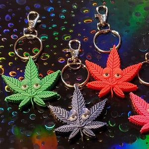 May include: A collection of cannabis leaf keychains in various colors, including green, red, orange, pink, and gray. Each keychain features a cartoon face and a silver clasp. The background is a dark, iridescent surface.