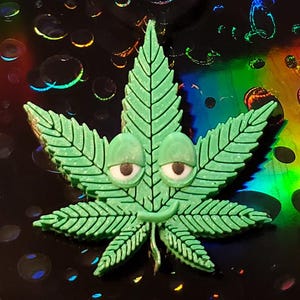 May include: A green cannabis leaf with cartoon eyes and a smile. The leaf is a vibrant green color with detailed veining. The background is a dark, iridescent surface with colorful, bubble-like patterns. The overall theme is whimsical and playful.